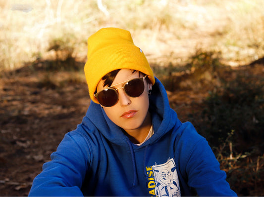Young person in a yellow beanie, blue hoodie, and round sunglasses sitting outdoors.