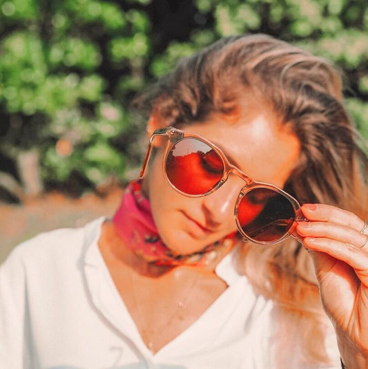 A woman tilts her head while adjusting orange-tinted sunglasses, wearing a pink neck scarf.