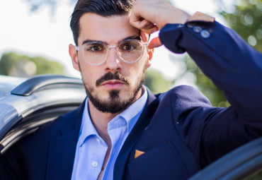A man with glasses and a beard, wearing a suit, stands near a car outside.