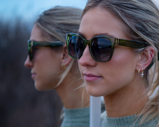 A woman in sunglasses stands next to her reflection in a mirror outdoors at dusk.