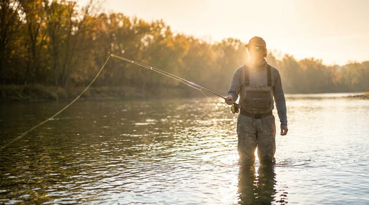 Angler wearing polarized sunglasses casting a fly rod while wading in a clear shallow river at sunrise