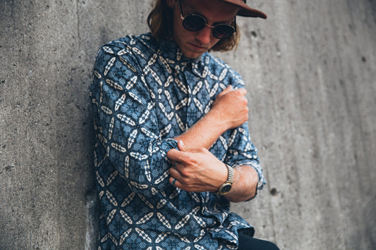 Man in a patterned blue shirt and sunglasses rolling up his sleeve against a concrete wall.